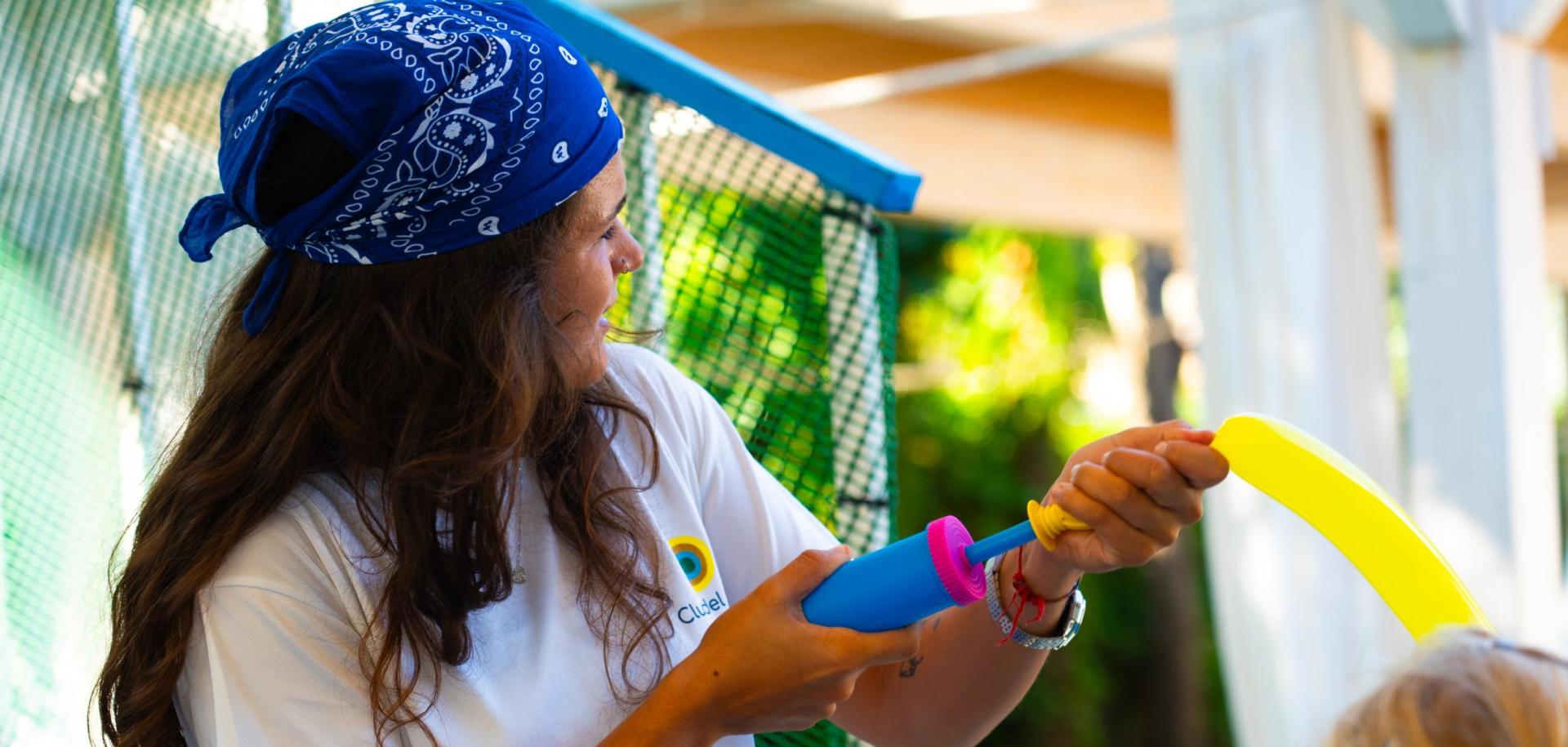 Persona gonfia un palloncino giallo con una pompa, indossa un foulard blu.